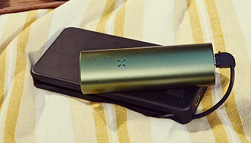 Chargement du vaporisateur Pax Flow Un vaporisateur pax flow vert élégant connecté à un chargeur portable noir via un câble usb, reposant sur une serviette à rayures jaune et blanche pour une charge pratique en déplacement.