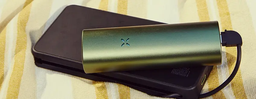 Chargement du vaporisateur Pax Flow Un vaporisateur Pax Flow vert élégant connecté à un chargeur portable noir via un câble USB, reposant sur une serviette à rayures jaune et blanche pour une charge pratique en déplacement.