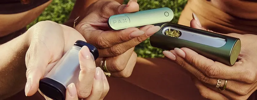 Remplissage du vaporisateur Pax Flow Deux mains collaborant pour remplir le vaporisateur Pax Flow avec du cannabis moulu provenant d'un petit grinder, illustrant le processus de chargement facile en extérieur sur de l'herbe verte.