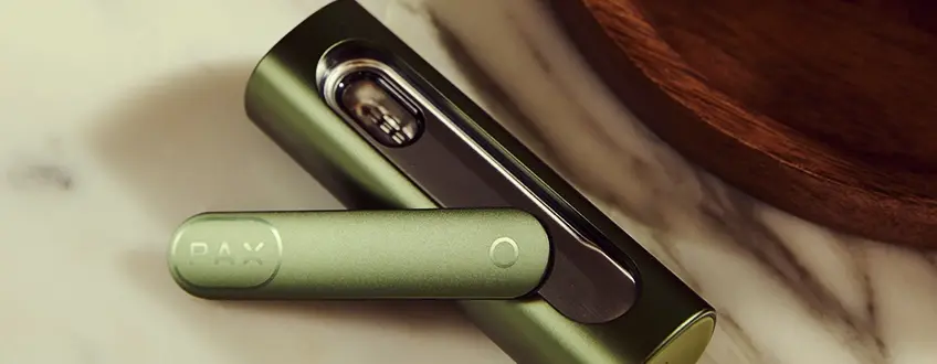 Vaporisateur Pax Flow ouvert Gros plan sur le vaporisateur Pax Flow ouvert révélant sa chambre interne remplie de cannabis, tenu par une main dans un cadre extérieur décontracté avec lumière naturelle.