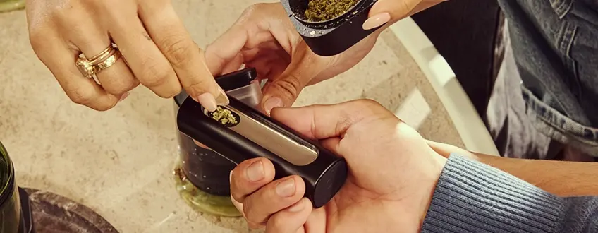Rechargement du vaporisateur Pax Flow Une main rechargeant la chambre du vaporisateur Pax Flow avec du cannabis moulu frais à l'aide d'une petite cuillère d'un grinder, posé sur une table en marbre pour un rechargement précis.