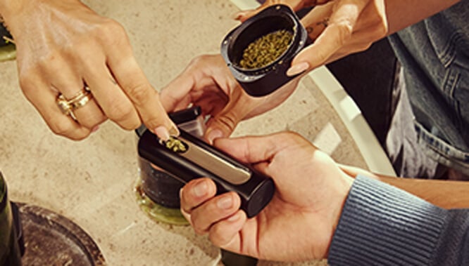 Rechargement du vaporisateur Pax Flow Une main rechargeant la chambre du vaporisateur Pax Flow avec du cannabis moulu frais à l'aide d'une petite cuillère d'un grinder, posé sur une table en marbre pour un rechargement précis.