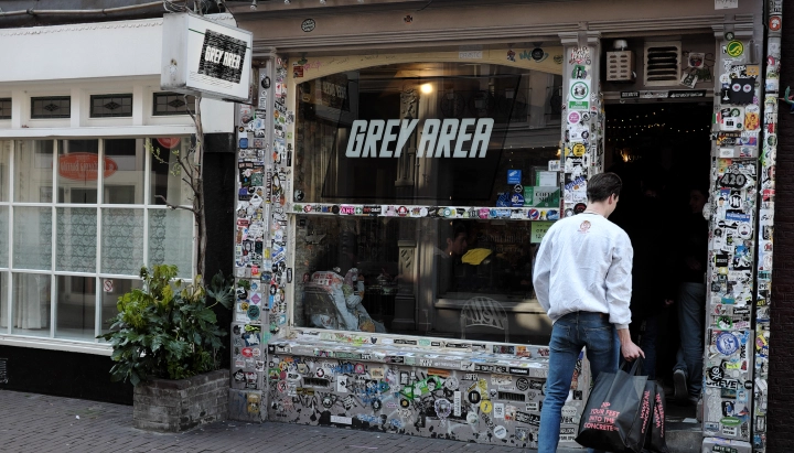 Entrée d’un coffeeshop de cannabis aux Pays-Bas avec une enseigne Grey Area et une devanture couverte d’autocollants.