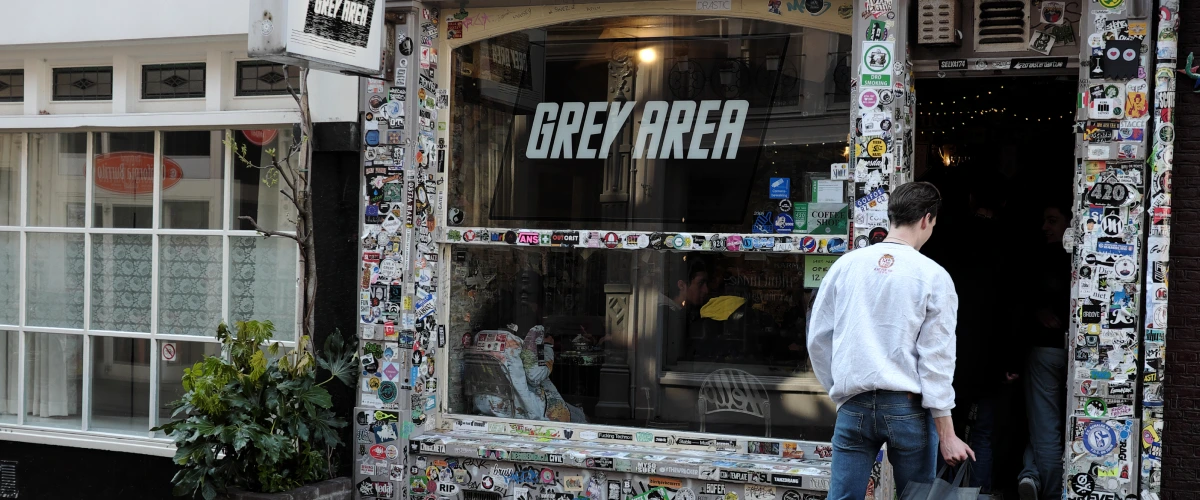 Entrée d’un coffeeshop de cannabis aux Pays-Bas avec une enseigne Grey Area et une devanture couverte d’autocollants.