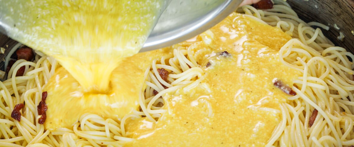 Mélange d’œufs et de fromage versé sur des spaghetti chauds dans une poêle, cinquième étape pour une carbonara infusée au cannabis